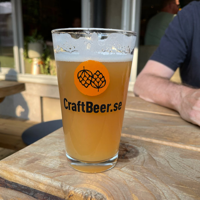 Ett glas oklar öl med logotypen CraftBeer.se, på ett träbord utomhus.