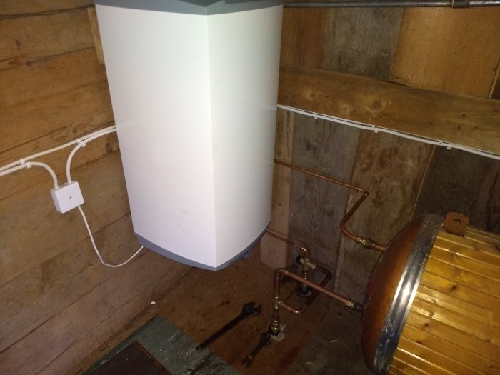 Nyinstallerad vit elberedare i ett trärums hörn med kopparrör och en gammal avstängd beredare.