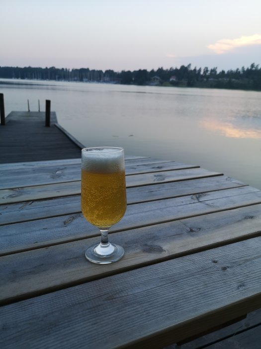 Ett glas öl på en träbrygga med en stilla sjö och skog i bakgrunden vid skymningen.