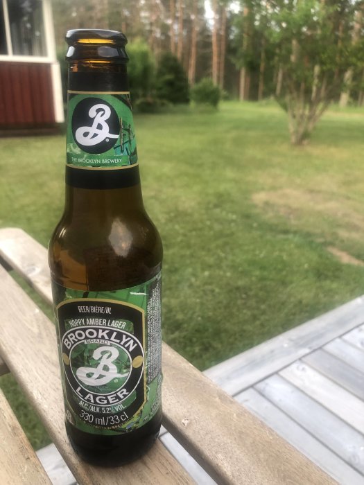 Flaska av Brooklyn Lager öl på ett trägeländer med en suddig trädgård i bakgrunden.