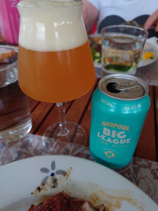 Ölglas fyllt med grumlig öl och en burk “Harpoon Big League Hazy DIPA” bredvid en maträtt på bord.