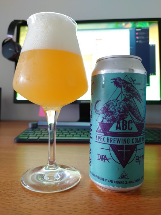 En bryggd öl i ett specialglas bredvid en burk från Apex Brewing framför en datorskärm.