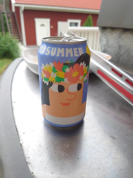 Ölburk med "Midsummer" design, placerad på en grå yta med röd stuga i bakgrunden.