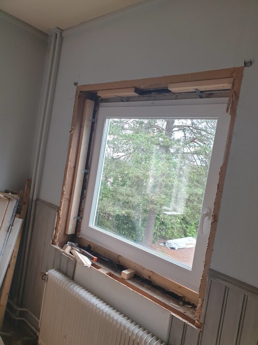 Renoveringsarbete av ett fönster med öppen vägg och synliga träbjälkar inomhus, med utsikt mot träd utomhus.
