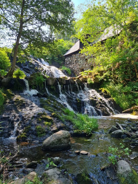 Vattenfall med stenhus och grönska under en blå himmel.