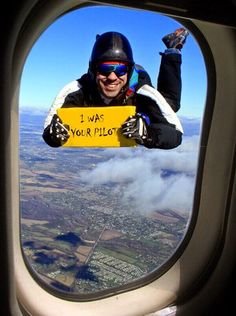 Skydivare håller en skylt med texten "I WAS YOUR PILOT" utanför ett flygplansfönster i luften.
