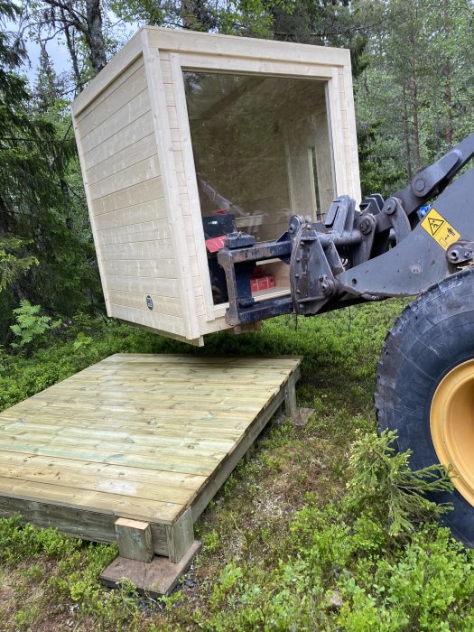 En ny bastu som lyfts av en hjullastare över ett nybyggt trädäck omgiven av skog.