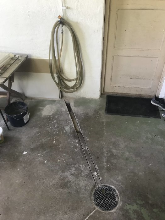Vattenkran med upprullad slang, spår kapad i betonggolv mot en gammal golvbrunn i ett garage.