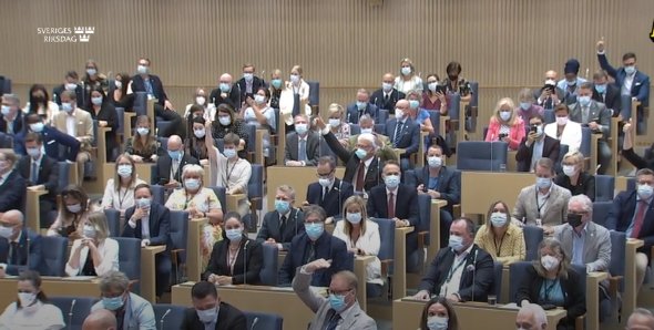 Människor i ett mötesrum, sittandes med munskydd, vid ett möte i riksdagen.