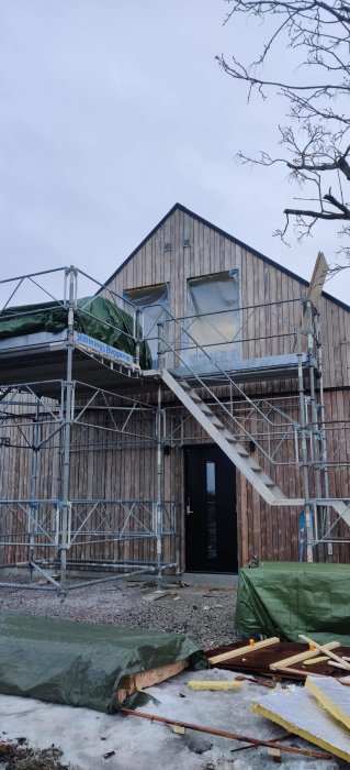 Byggnadsställning vid 1,5-plans trähus under renovering, med oöppnade fönster och byggmaterial på marken.