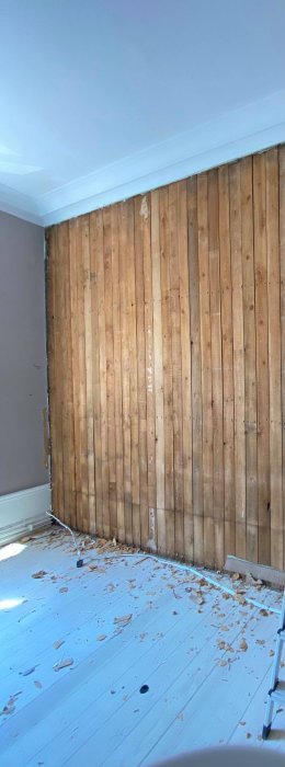 Orenoverad vägg med lodräta träplank och spill på trägolv i sekelskifteslägenhet.