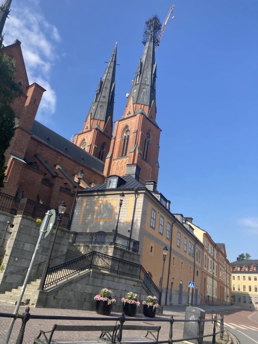 Röd tegelkyrka med två höga torn och en byggkran, omgiven av stenlagda trappor och en gatlykta.