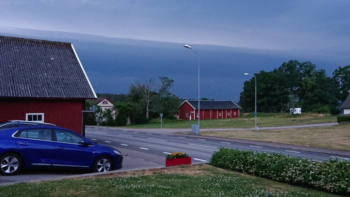 Mörka moln över en lantlig väg med en blå bil, röda hus och en blomlåda.