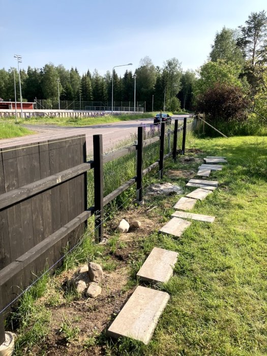 Svartmålat staket med tryckta stolpar och tvärliggare, bredvid väg och grönt gräs, med plattgång längs med.