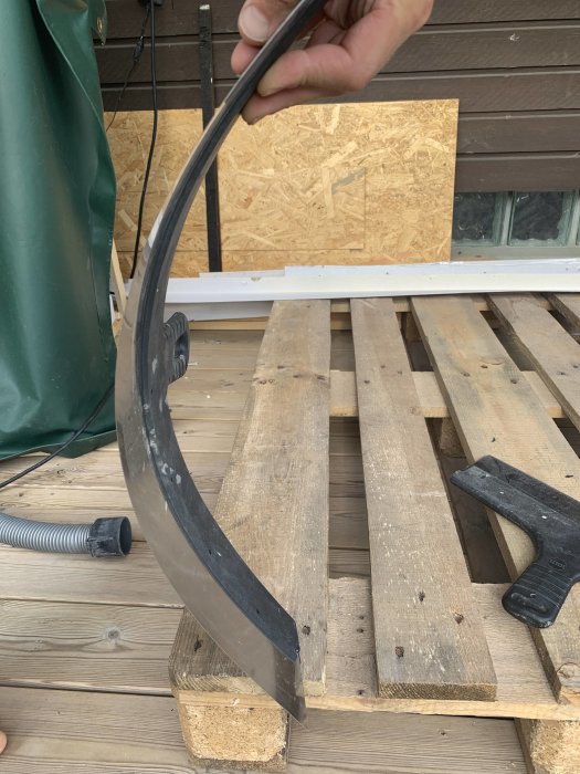 Hand håller upp en böjd skackelspade framför en träpall, bredvid en gummihammare.