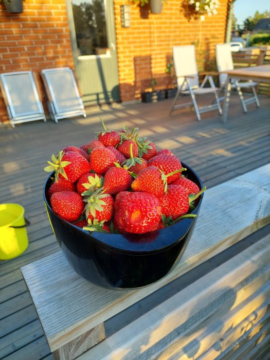 En skål med mogna jordgubbar på ett träbord på en terrass, redo att skördas.