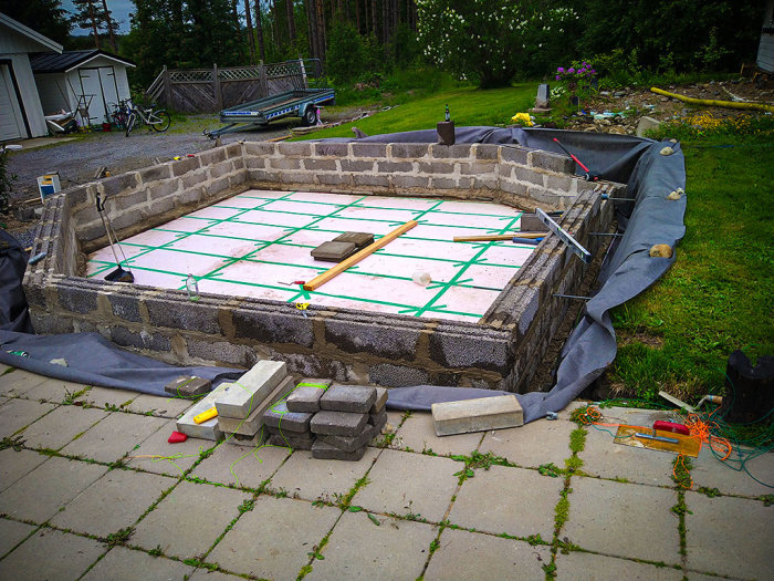 Grund för rund pool under konstruktion med murade stenblock och armering på plats, i en trädgård.