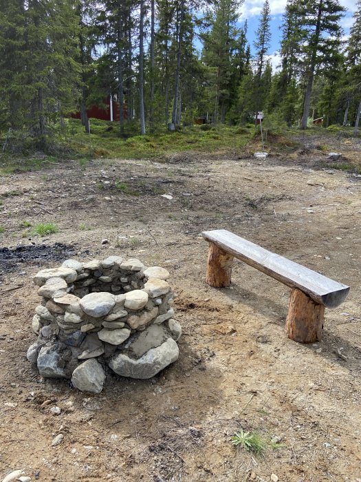 En handbyggd eldstad av sten och en träbänk utomhus i en skogsglänta.