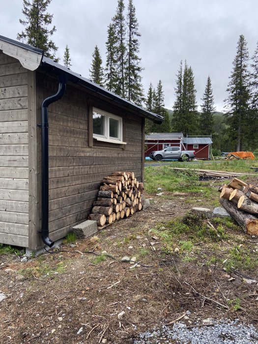 Uppstaplad ved vid stuga med skog i bakgrunden, halvfärdigt utomhusprojekt.