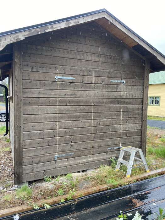 Träförråd med pardörrar och en liten stege framför, omgiven av gräs och asfalterad väg.