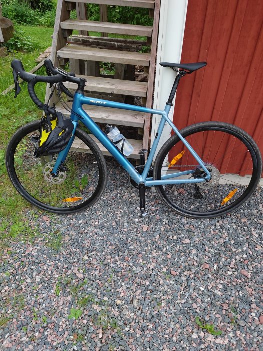 Blå landsvägscykel lutad mot en trätrappa bredvid en röd stuga, står på en grusgång.