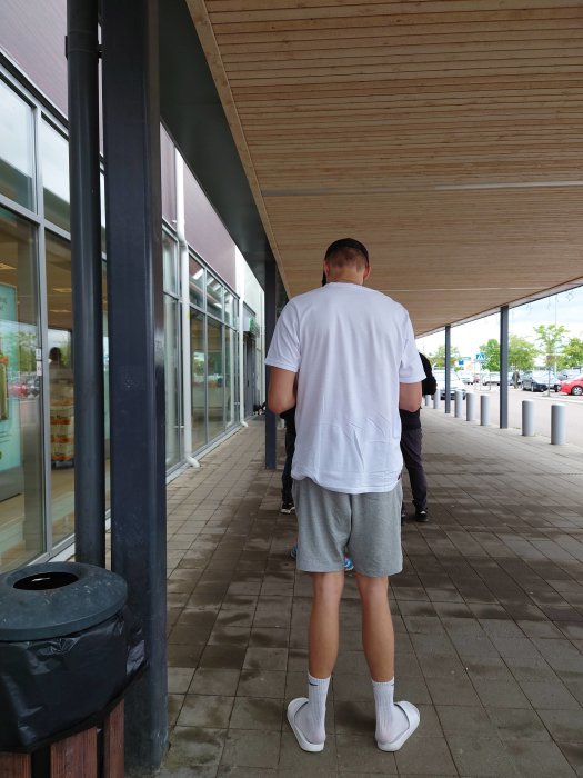 Person i vitt står framför en byggnad med stora fönster under ett träbeklätt överhäng.