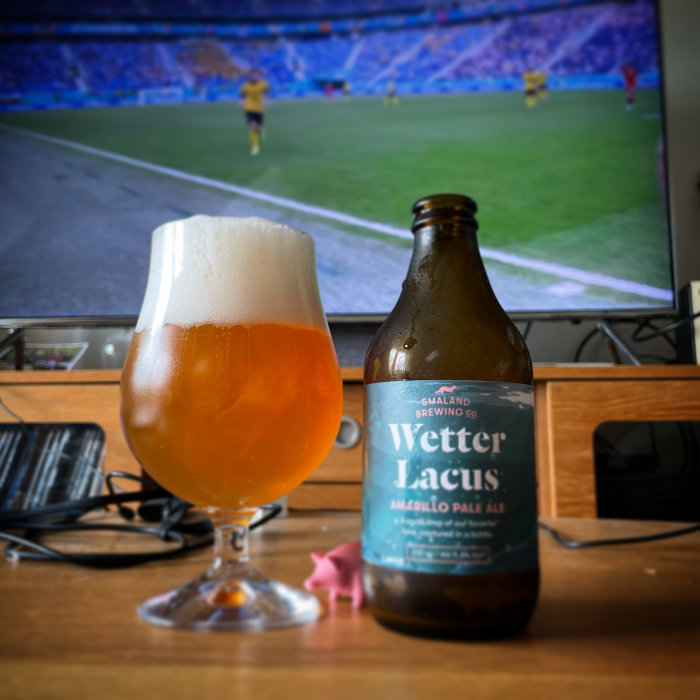 Ölglas och flaska med etikett "Småland Brewing Co. Wetter Lacus" framför fotbollsmatch på TV.