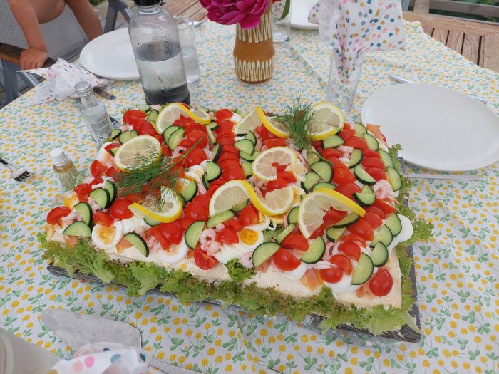 En smörgåstårta dekorerad med räkor, ägg, gurka, tomat och citronskivor på ett bord.