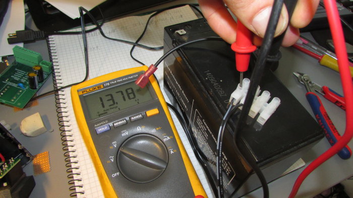 Multimeter visar 13,78 V under test av modifierad batteriladdare på arbetsbord fyllt med elektronikdelar.