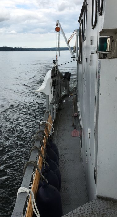 Sida av ett skepp med förtöjningsfendrar hängande längs relingen, utkik över sjö.