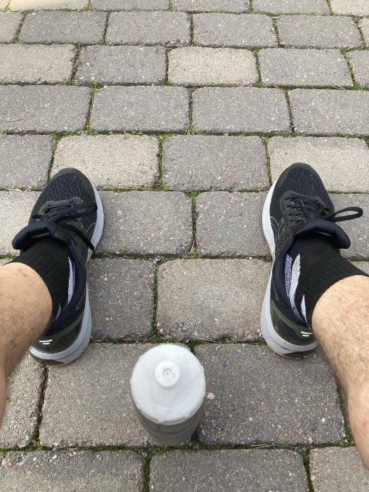 Person sitter på marken med löparskor, ben synliga, framför en grå plasthantel på stenläggning.