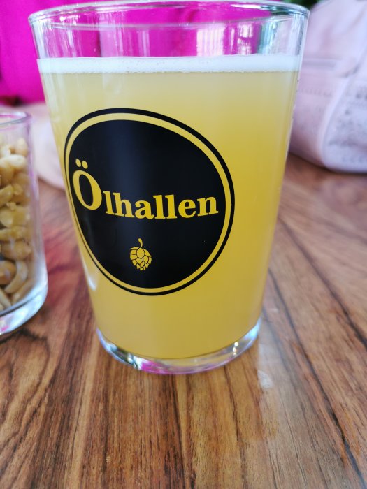 Ett glas med gul öl märkt "Ölhallen" på ett träbord med en skål jordnötter i bakgrunden.