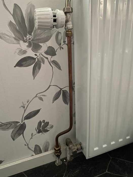 Radiator med termostat ovanpå och röranslutningar nedtill mot blommig tapet.