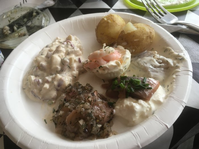 Paper plate with potatoes, shrimp salad, pickled herring, and creamy sauce on a patterned tablecloth.