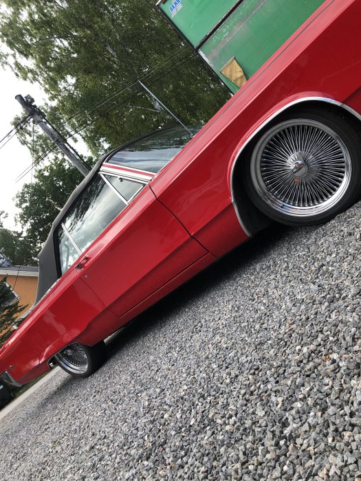 Röd lågprofilbil med ekersfälgar parkerad på en grusyta, snedvinkel.