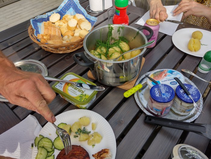 Sommarlunch på trädäck med nya potatisar, sill, bröd, gräddfil och en öppnad ostförpackning.