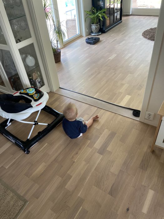 Barn sitter på trägolv och tittar på en robotdammsugare i ett hem.