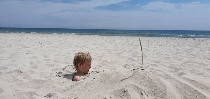 Barn ler och sticker upp huvudet ur sanden på en strand med havet i bakgrunden.
