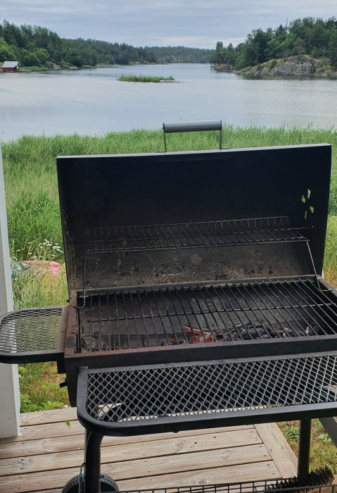 Grill vid sjökant med öppet lock, aska på grillgallret, natur och vatten i bakgrunden.