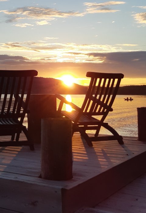 Solnedgång vid vattnet med silhuetter av två trästolar på en brygga och en kanot.