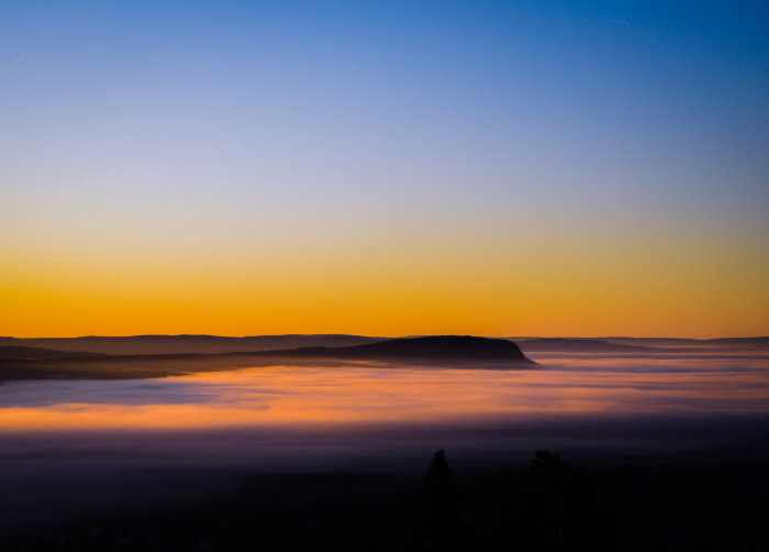 Soluppgång med ljusa himmelstoner över ett kuperat landskap och dimtäckta dalar vid tidig gryning.
