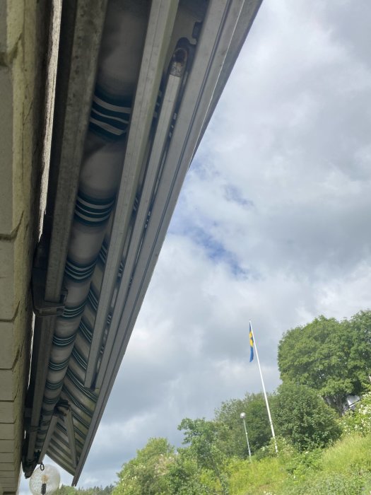 Markis som är delvis indragen med synliga mekaniska delar och en svensk flagga i bakgrunden.