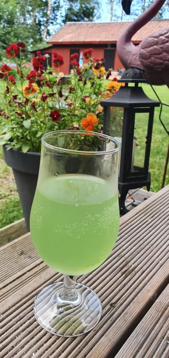 Ett glas med en grön dryck på ett träbord med suddig bakgrund av blommor och en röd stuga.