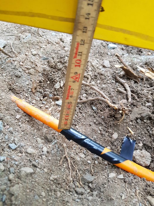 Måttband som visar grävdjupet för en skadad orange kabel omgiven av jord och småsten.