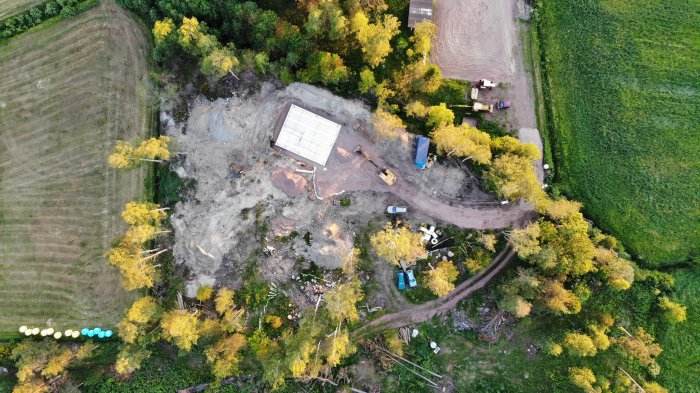 Drönarvy av byggplats med delvis uppförd byggnad omgiven av arbetsfordon och natur.