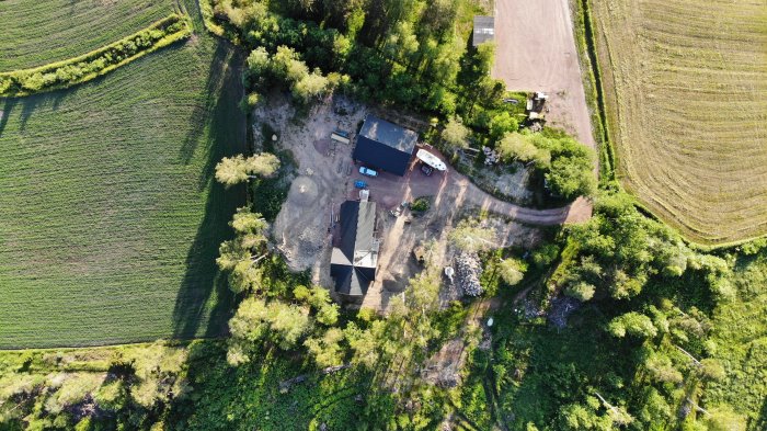 Flygbild över byggprojekt med halvfärdigt hus och takarbete, omgivet av gröna fält och skog.