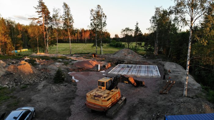 En grävmaskin på en byggplats vid ett pågående byggprojekt av ett hus med skog i bakgrunden i skymningen.