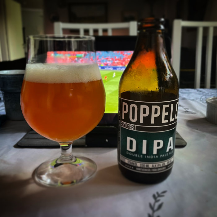 Ölglas med öl vid sidan av en flaska märkt 'Poppels DIPA' framför en TV som visar fotboll.