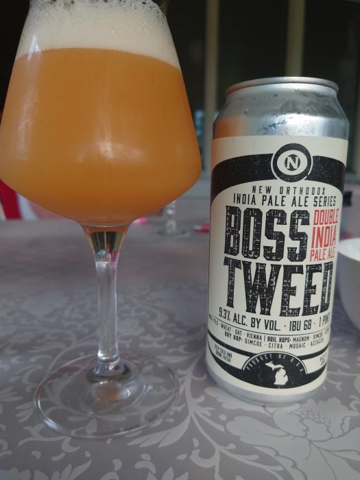 Glas med öl bredvid en burk märkt "Boss Tweed Double India Pale Ale" på ett mönstrat bord.