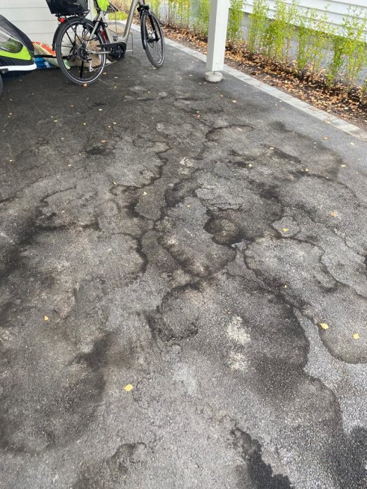 Fläckig och flammig asfalt under carport med synliga brungråa ringar och en cykel parkerad i kanten.